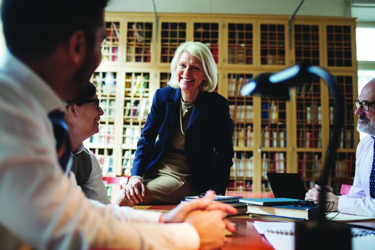 A person half-sitting on a table, smiling at others seated. A large bookcase behind them.