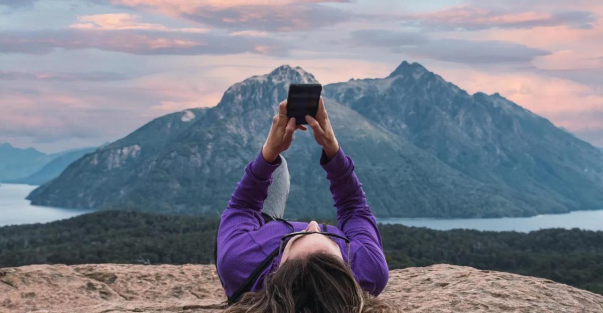 A person laying on a rock looking at their phone held above them. A mountain landscape in the distance.