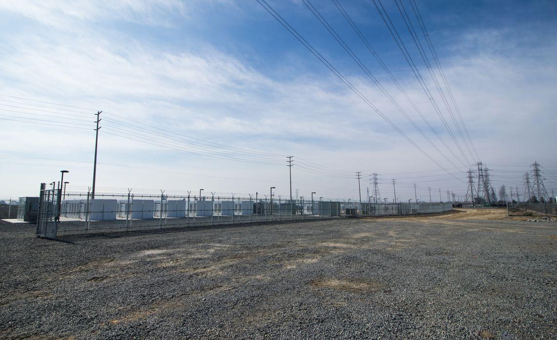  A power station with tall power lines above and graveled pad in front.