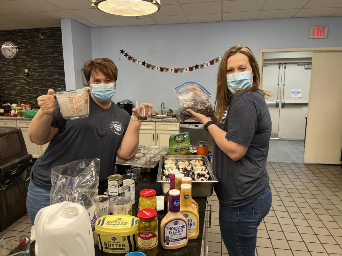 Two Bath & Body Works associates stand at a counter in the Ronald McDonald House kitchen. One associate is holding up a measuring cup filled with yogurt and a small cup of blueberries. The other associate is holding up a bag of blueberries. On the counter are small cups filled with yogurt and topped with fresh fruit.