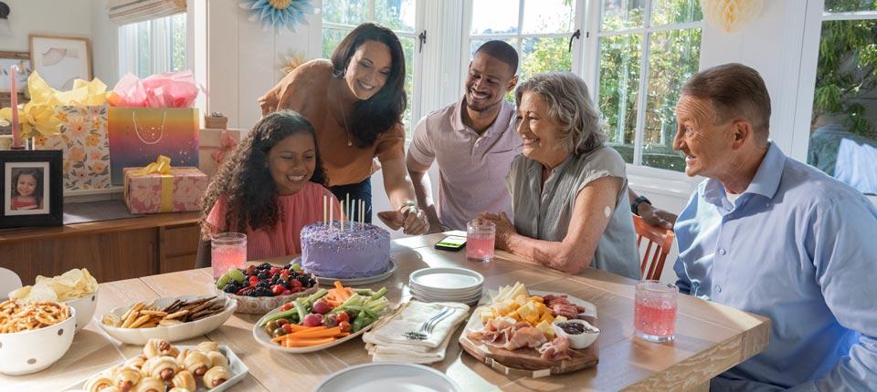 People sat around a buffet table, looking at a person who is sat in front of a celebratation cake