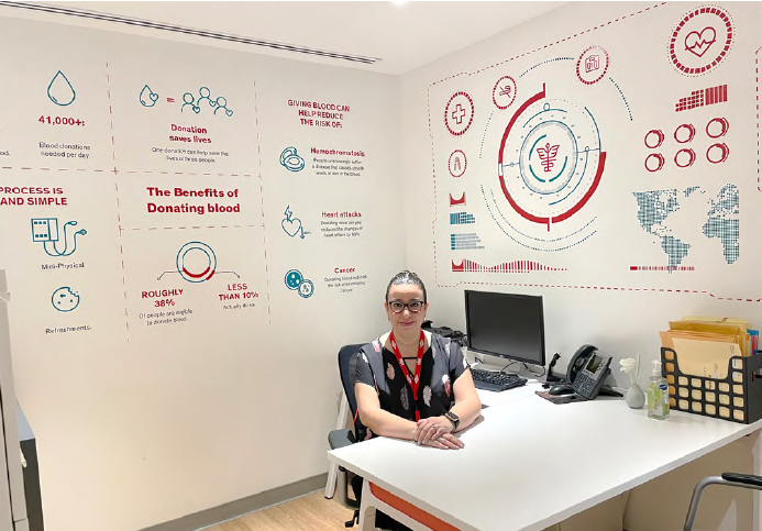 A person sitting at a desk in an office. A large informative mural on the wayy "The benefits of donating blood"