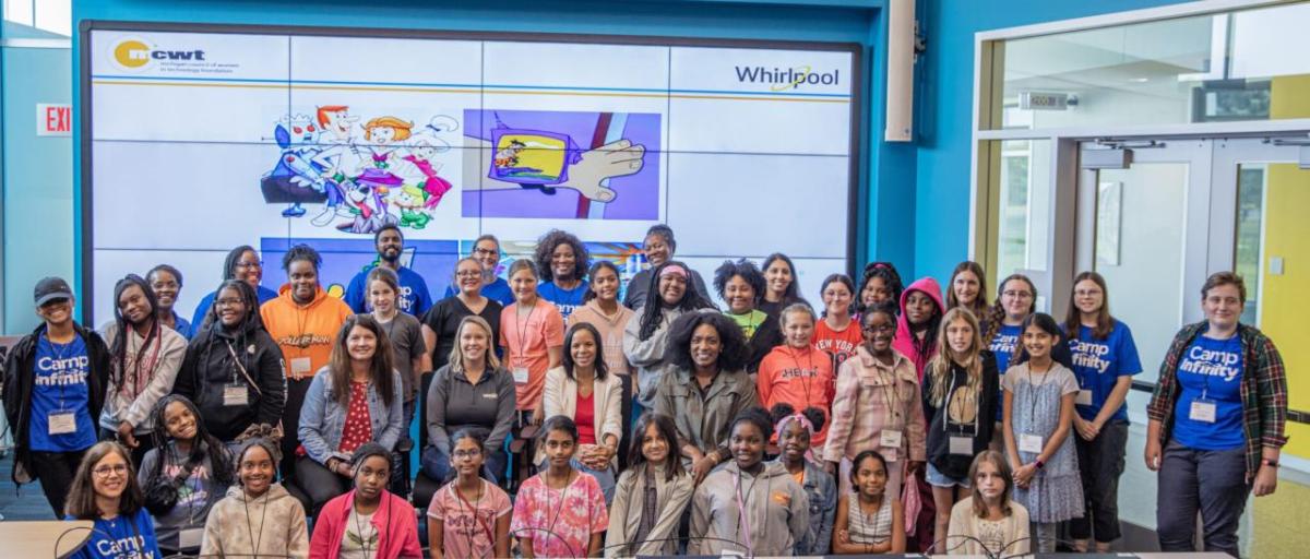 A group of children and adults posed in a classroom with a large digital screen behind them.