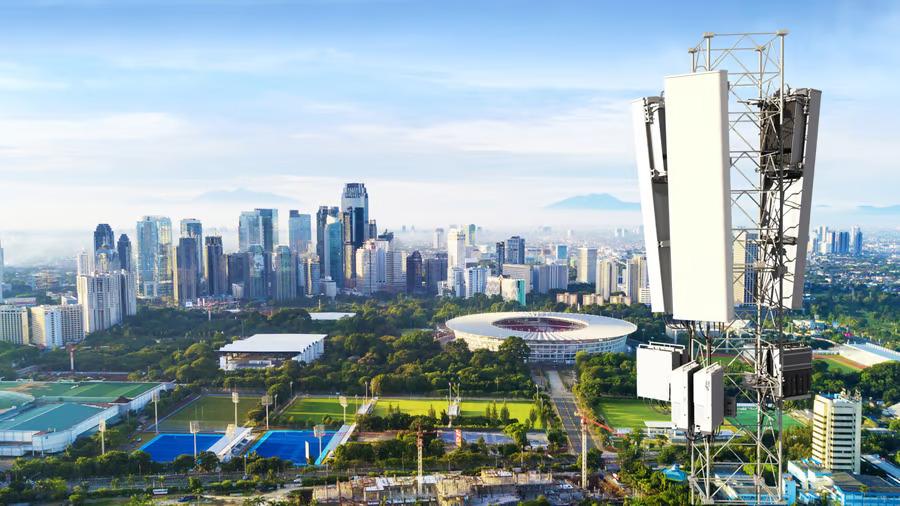 Elevated view of the top of a cell tower with a city skyline and urban area behind it.