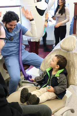 A dentist plays with a child in a dentist chair, a tooth mascot in the corner, others around smiling.