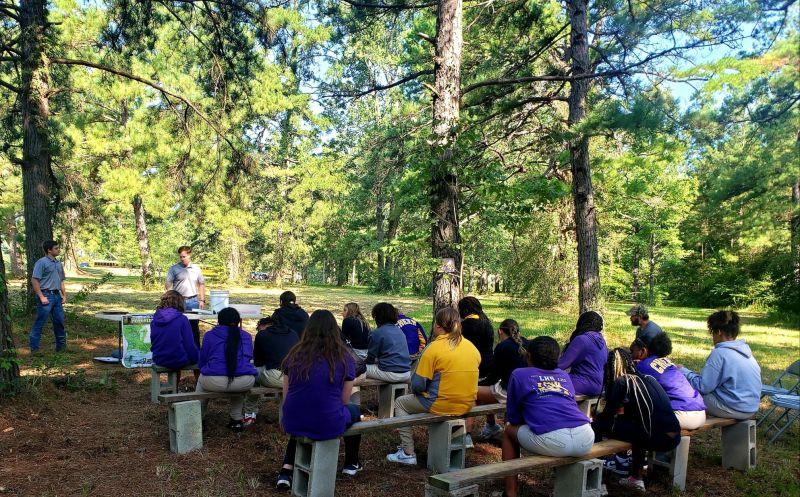 Students sitting on benches in a park setting watch a speaker