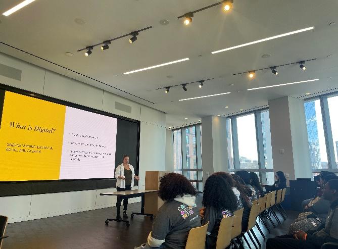 A classroom of students, a presenter at the front with digital display behind them.