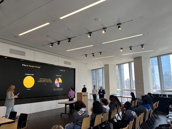 A classroom of students, multiple presenters at the front and digital display of pie charts behind them.