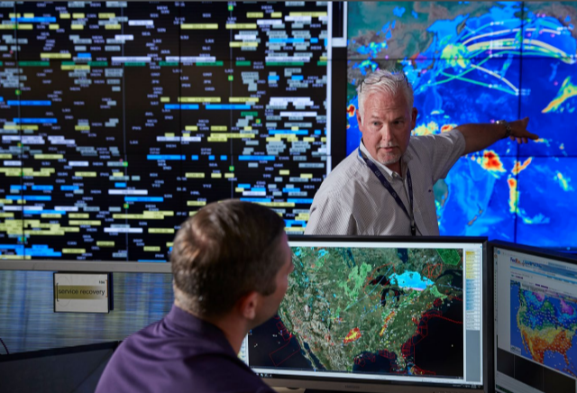 in a room with large screens on the walls, a person points at a global map while talking to a seated person looking at a computer screen with a map of the US