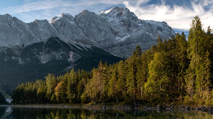 Landscape of a forested shore of a body of water, snow capped mountains in the distance.