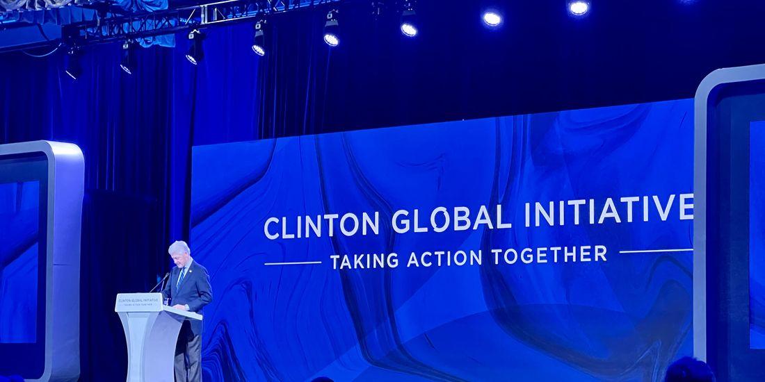 Bill Clinton at a podium, a large screen with "Clinton Global Initiative" behind him