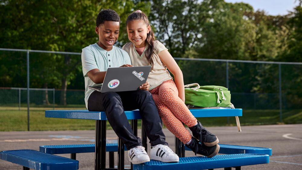 Two smiling children looking at an open laptop while sitting on an outdoor table.