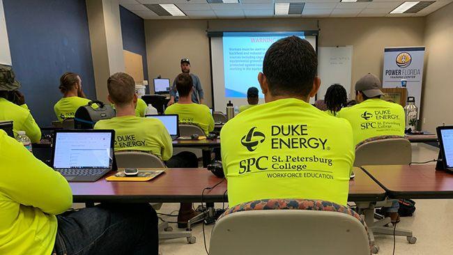 Students in matching shirts in a classroom setting, a person speaking at the front of the room.