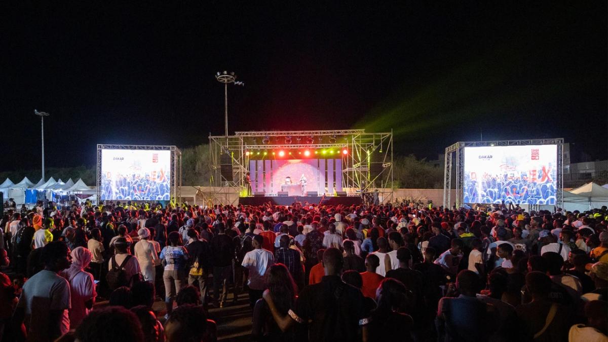 A large event with many people standing, looking at the stage and projected screens on each side. Performers on stage.