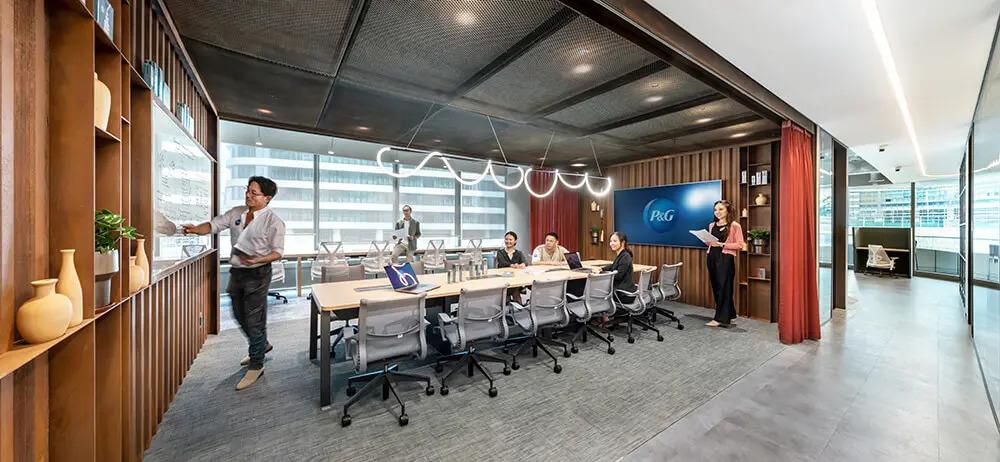 People seated at a long table with rolling office chairs, a long open wall, and person at a board near the front of the space.