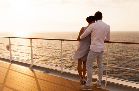a couple hugging, leaning against the rail of a cruise ship. Setting sun in the background.