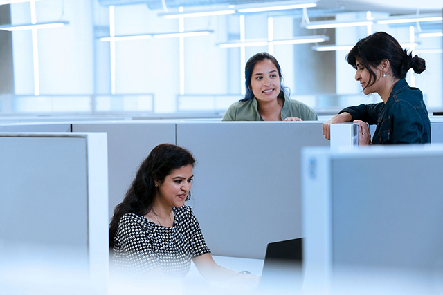 Two people standing, talking with a third who is seated at a desk in a cubicle, working on a laptop.