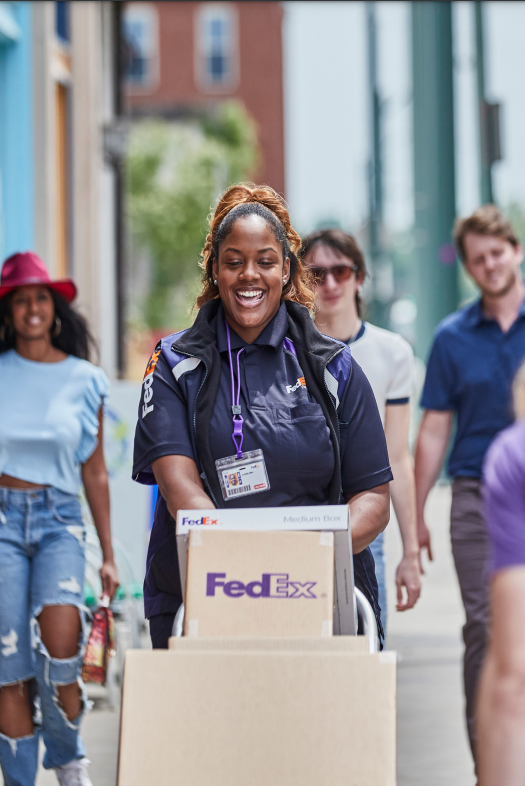 A fedex delivery person with a cart of boxes, people walking behind them. 