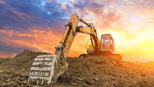A large construction digger on a hill of dirt, a bright setting sun behind it.