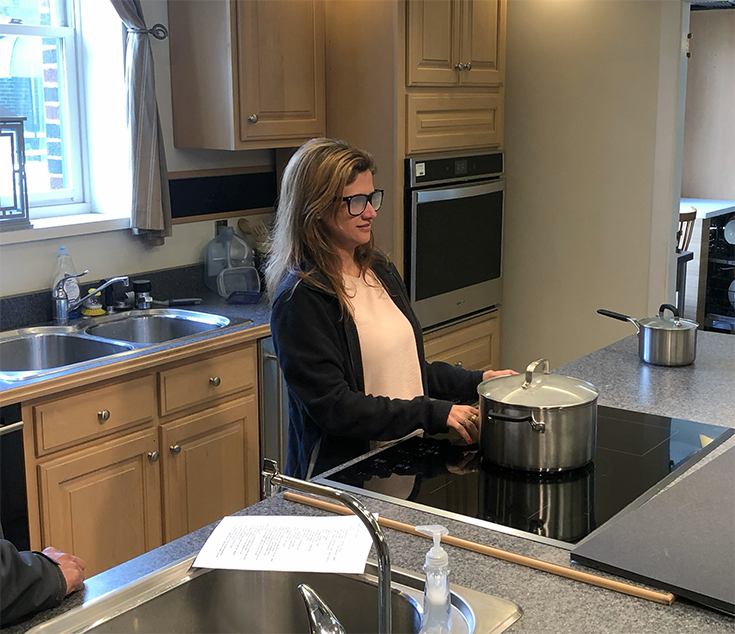 woman wearing frosted glasses stands at an induction range and touches a silver pot with the back of her hand