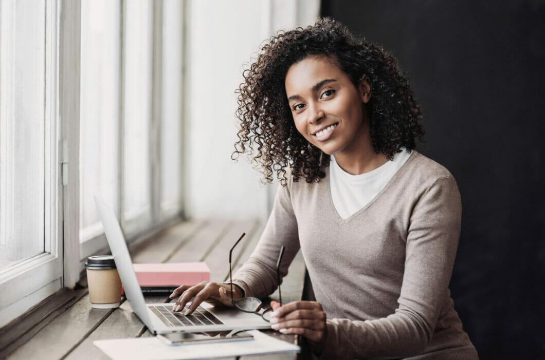 Photo 198117357 / Social© Poike2017 | Dreamstime.com Young woman sitting at a desk, looking at the camera and typing on her laptop.
