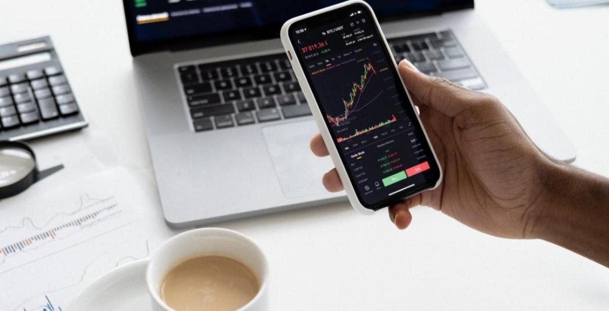 Photo courtesy of Nasdaq.com A hand holds a smartphone, with a cup of coffee and a laptop shown on a desk