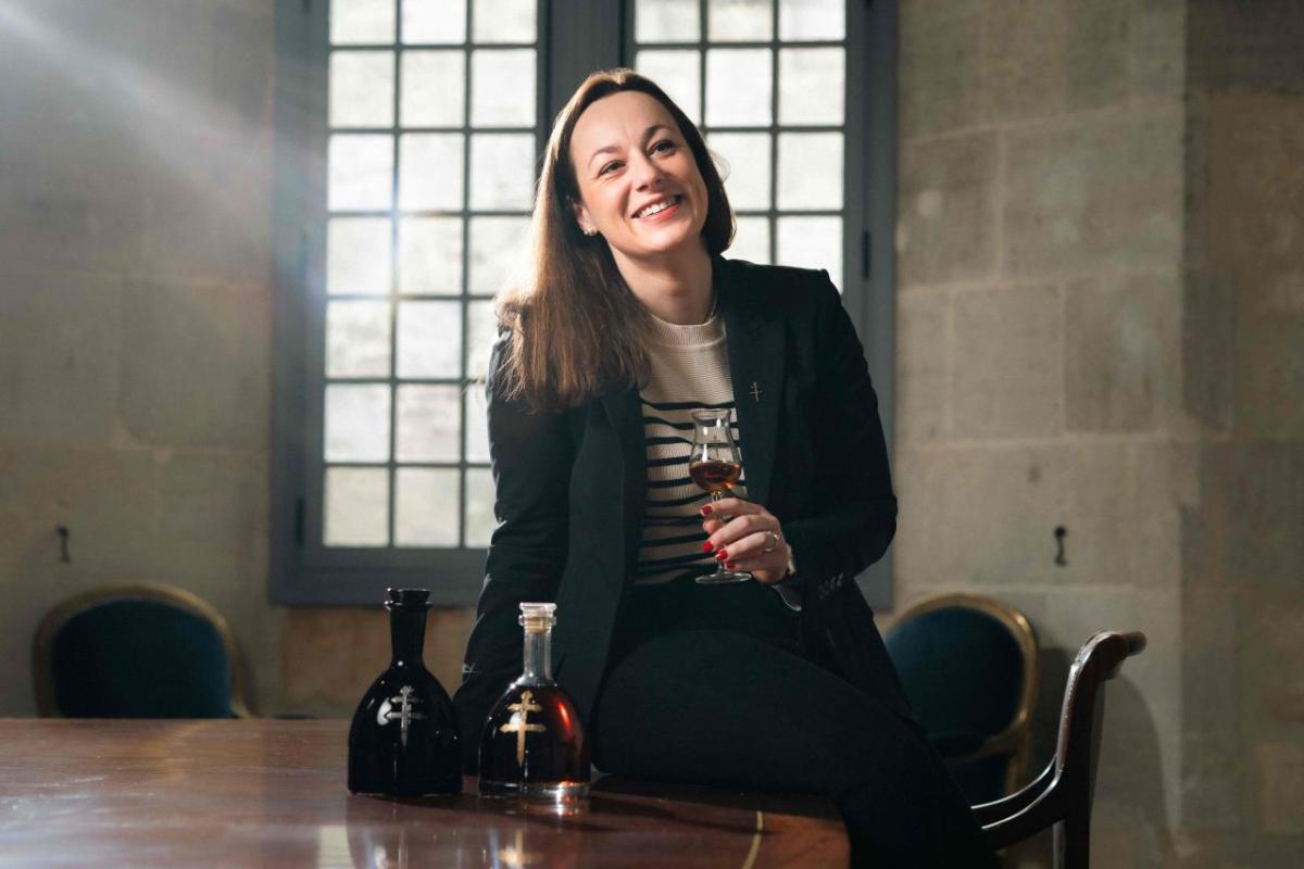 Agathe Boinot half-seated on a table. Bottles of cognac next to her. A small glass in her hand.