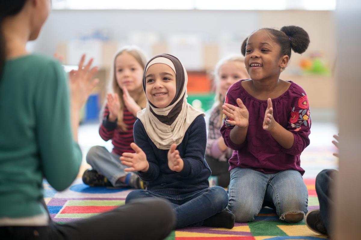 children sitting on a colorful floor watch and adult and are clapping their hands