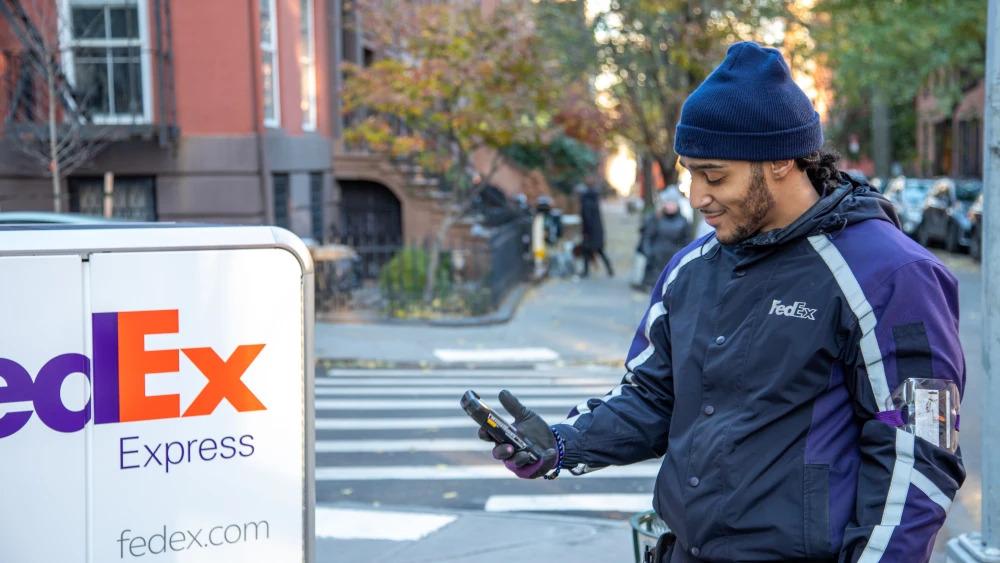 A person in FedEx uniform next to a tall cart with FedEx Express logo on it. They are looking at a hand-held device on a sidewalk in a residential area.