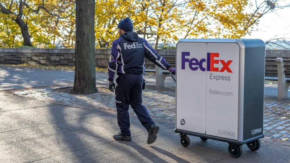 A person in FedEx uniform pulling a tall cart with FedEx Express logo on it through a park setting.