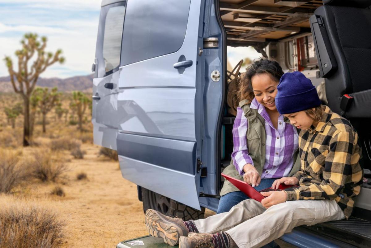 An adult and hild looking at an electronic tablet while sitting in the open side of a van in an arid area.