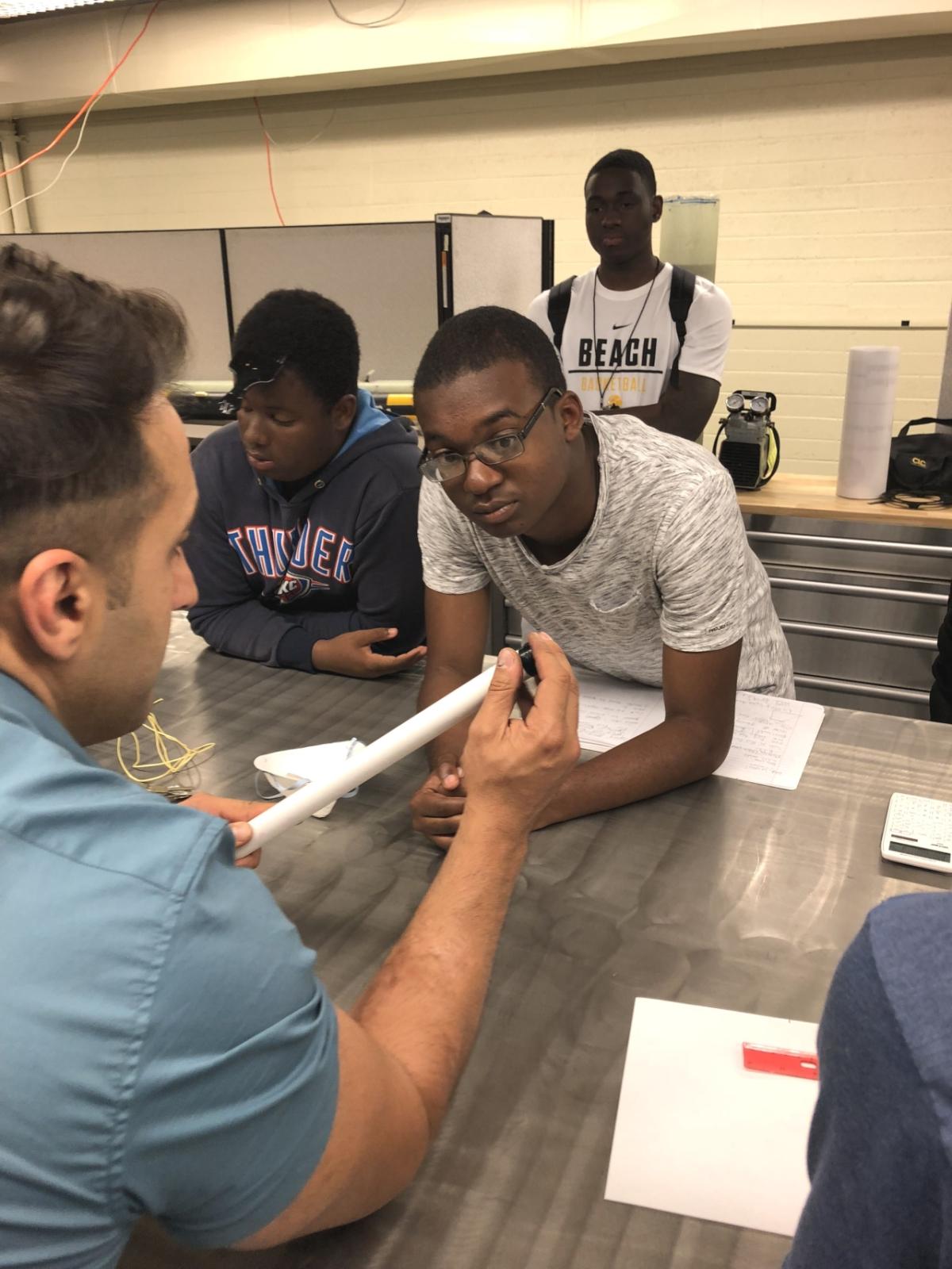 Three students watch an instructor assemble a piece of equipment