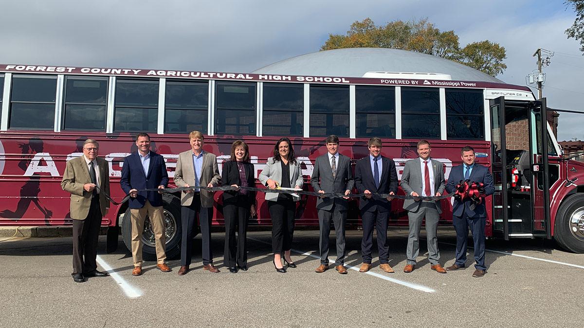 A row of people holding a ribbon in front of a school bus.