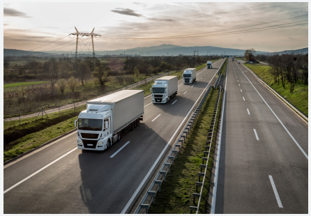 A line of semi trucks going down a road, wind turbines in the background.