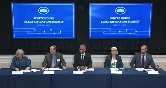 Five people sitting at a long desk, Two screens behind them with "White house Electrification summit" on them.