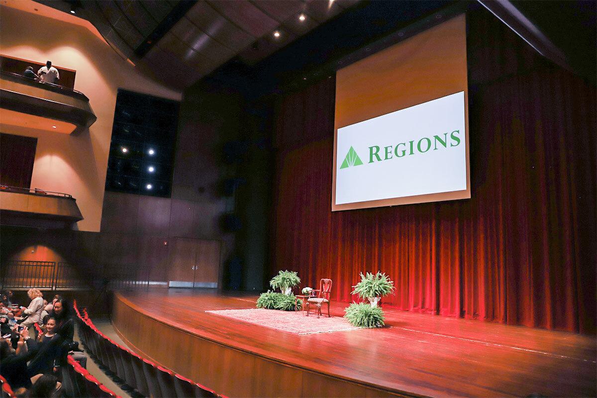 an empty stage with a chair and greenery. a large screen with Regions on it