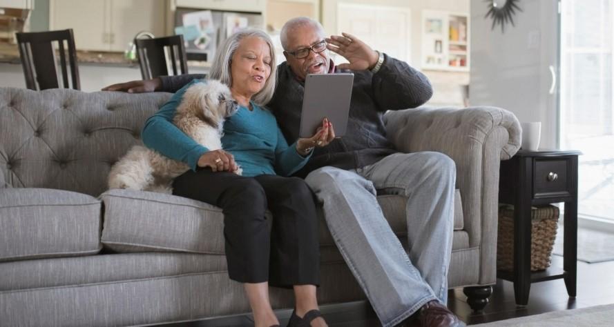 Two people and a small white dog sitting on a couch, looking at a tablet device.