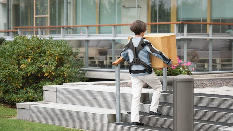 a petite person wearing an 'exoskeleton' carrying a box up steps