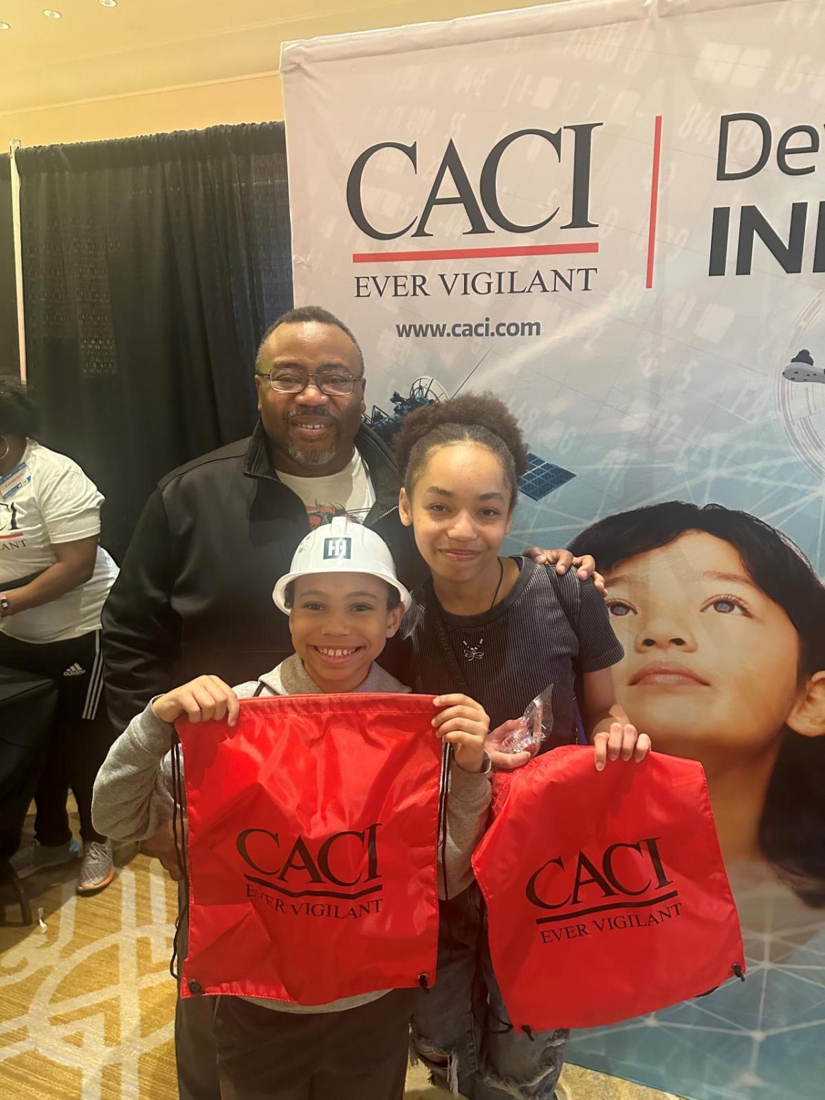 A group of three pose in front of a CACI banner, holding a CACI bag.