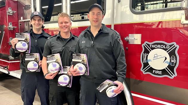 Firefighters at Thorold Fire in Thorold, ON display just a few of the 76,000 combination smoke and CO alarms installed across Ontario since 2009 as part of Safe Community Project Zero. Three firefighters holding new carbon monoxide detectors, a fire truck behind them.