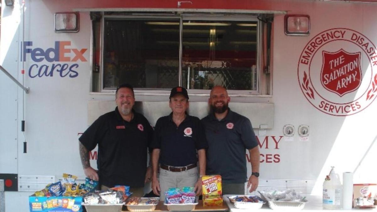 Three people standing in front of a box truck with a table of food in front.