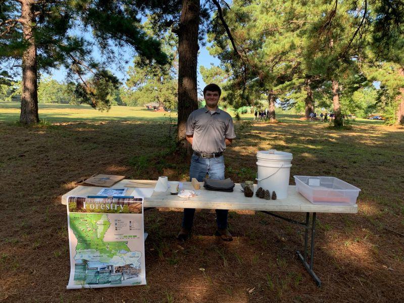 A person standing in a park behind a table with presentation materials and a map of Louisiana.