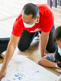 An adult kneeling on the floor with children, coloring on a piece of paper.