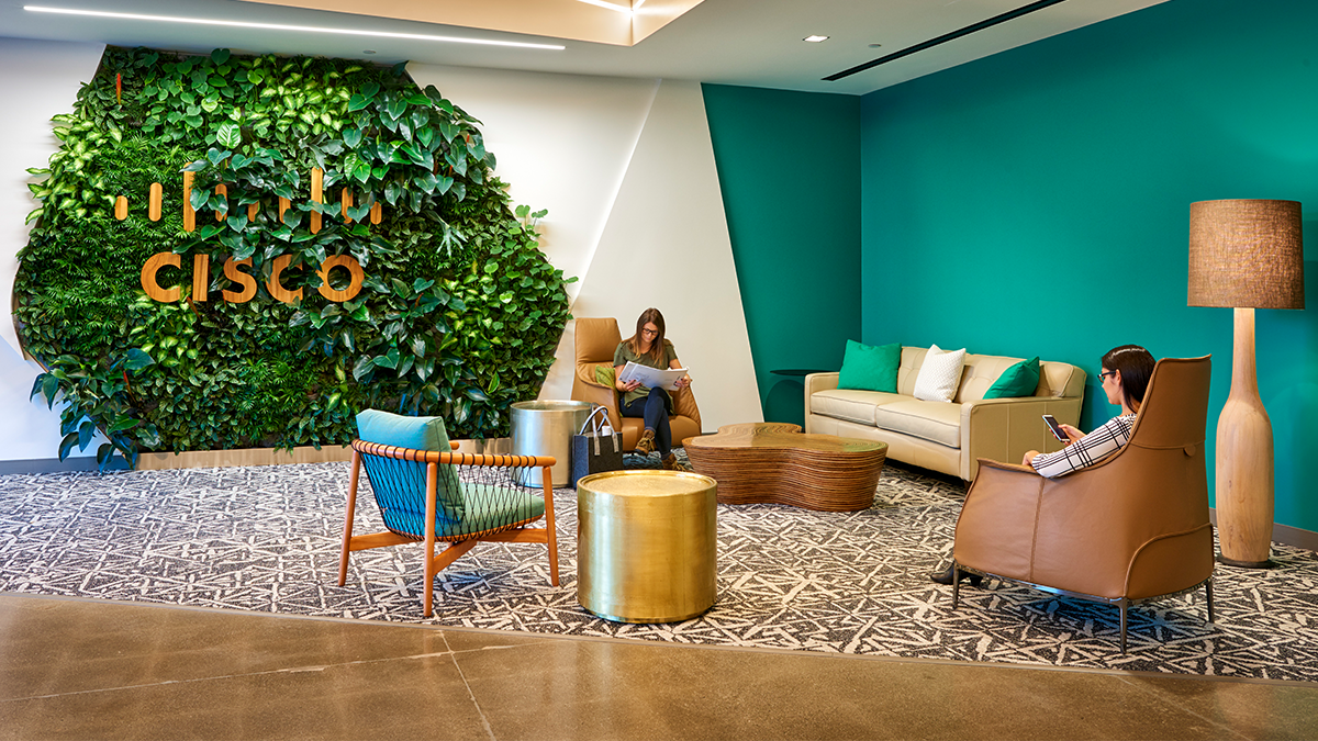 people sitting in a living room setting, a wall of greenery behind them with Cisco logo on it