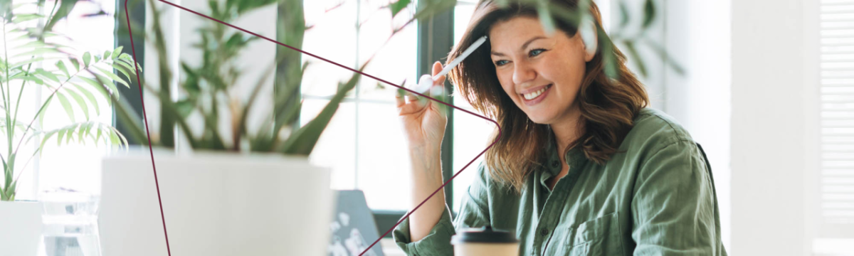 A smiling person looking at a laptop, a pen by their head.