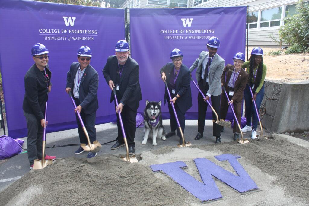 seven people in hard hats holding shovels dug in to sand around a large "W" and one very good dog in the middle