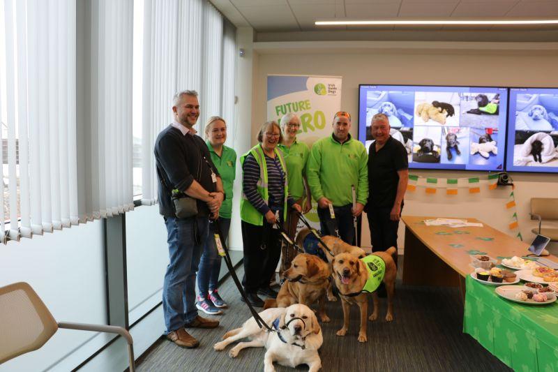 A group of six people and their guide dogs in an office room with food on the tables. All good dogs.