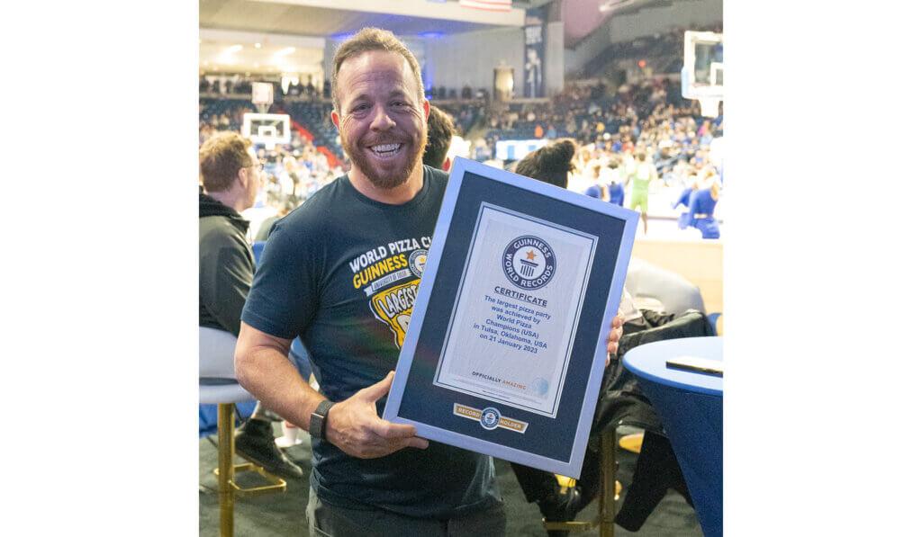 A person holding an official, framed, certificate from Guinness world records. A crowded stadium and basketball court behind them.