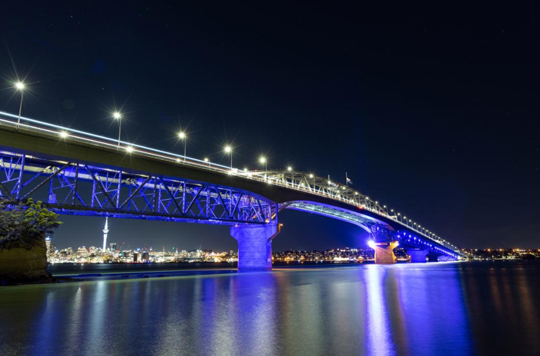 night panorama of Auckland Harbour Bridge over an expanse of water and is lit up in blue lights