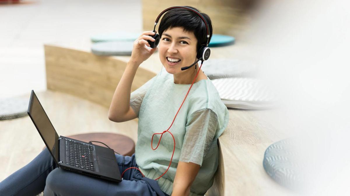 A person smiling at the camera, lifts a side of the headphones they're wearing.